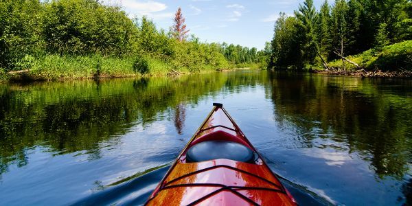 The bow of a kayak on a waterway bordered by green trees